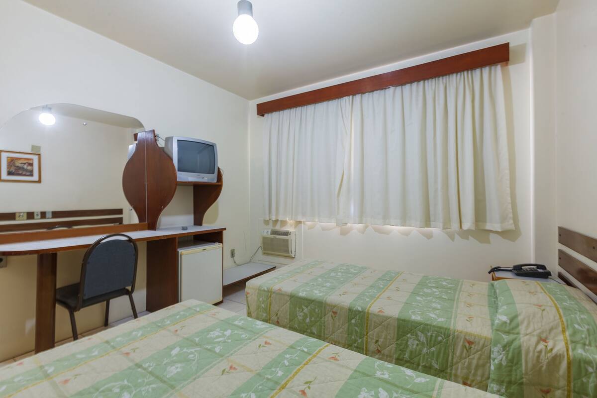 A room with two beds, a desk, a chair, a television, and a window with curtains.