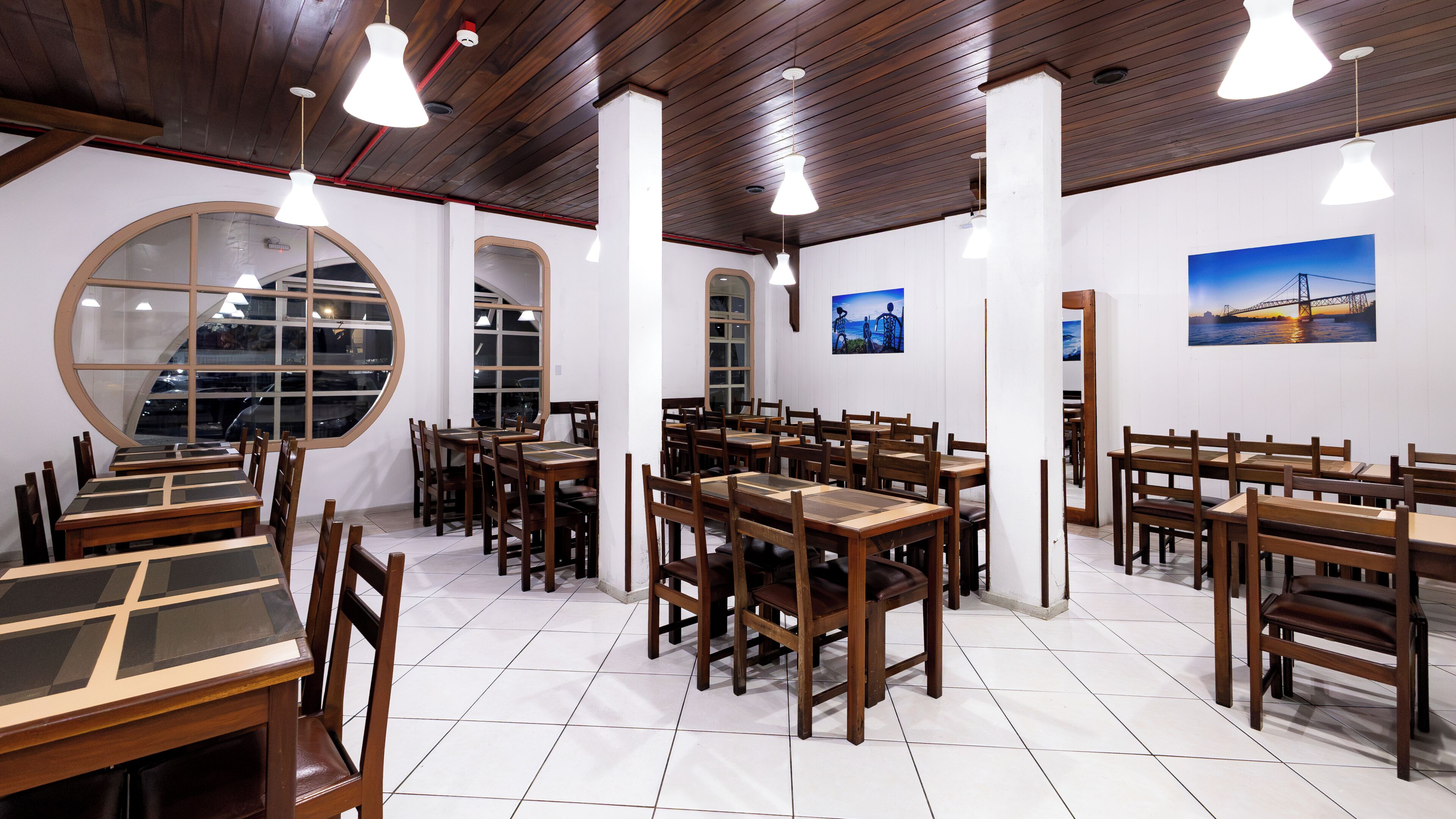 Un restaurante con mesas y sillas de madera, ventanas amplias y lámparas colgantes.