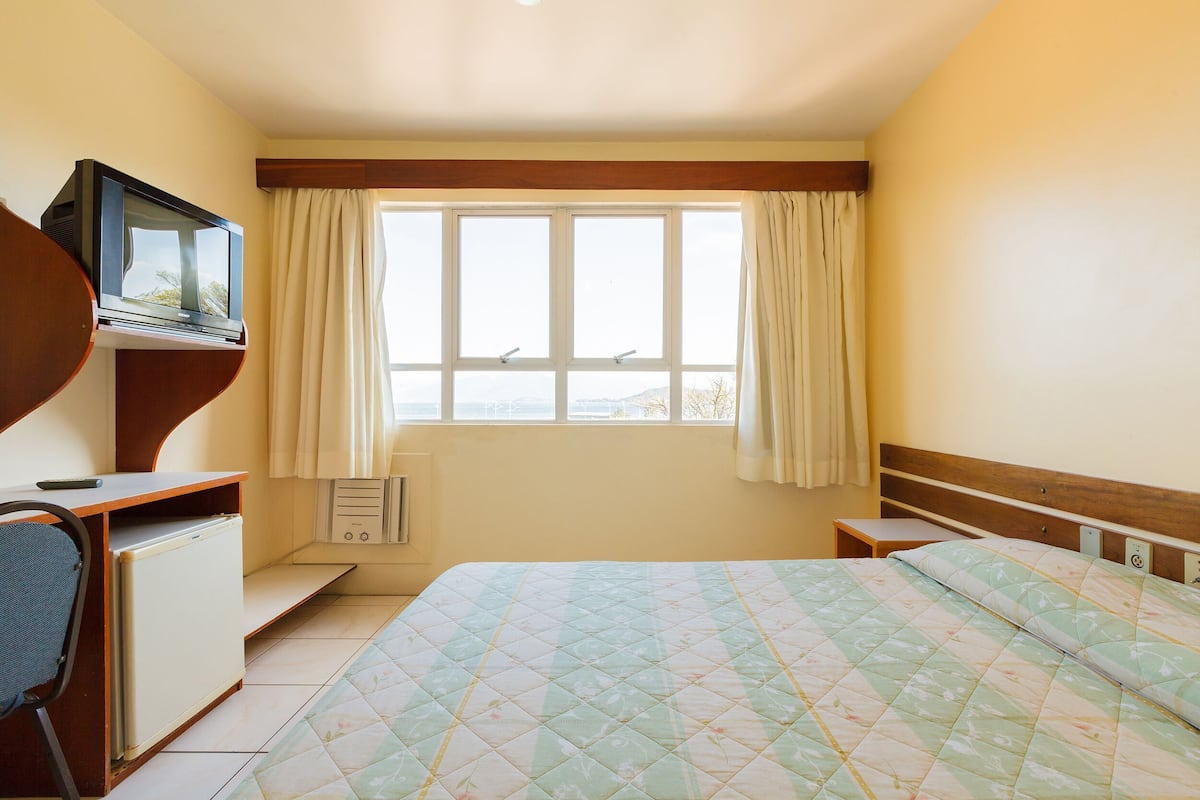 A bedroom with a bed, a TV on a stand, a desk with a chair, and a window with curtains.