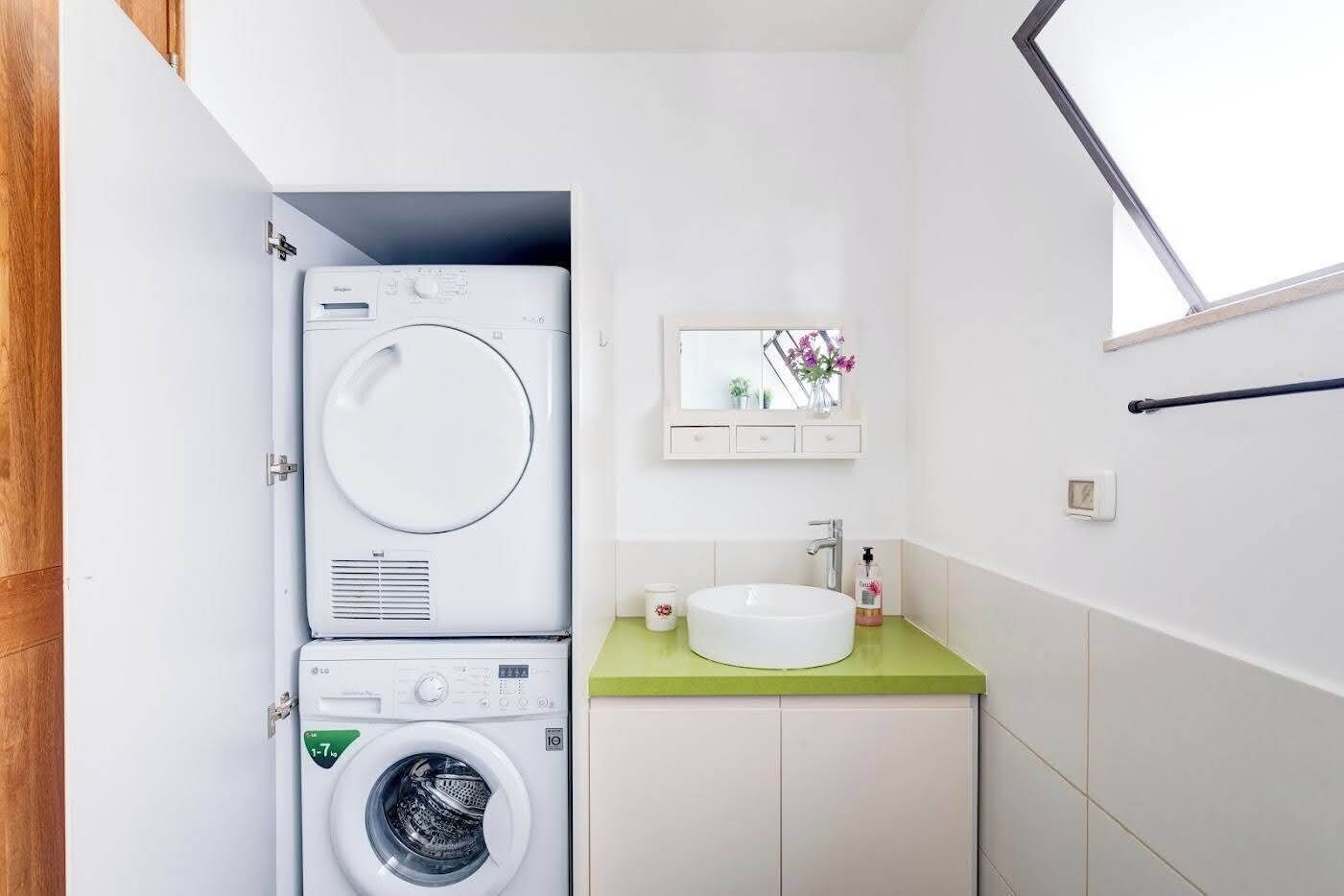 Un lavadero pequeño con lavadora y secadora apiladas, un lavabo con encimera verde y una pequeña ventana sobre el espejo.