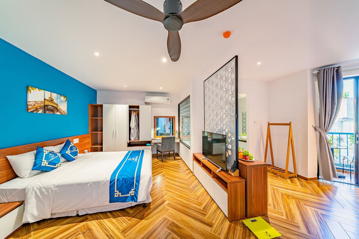 A modern hotel room with a bed, TV, wooden flooring, and a balcony with curtains.