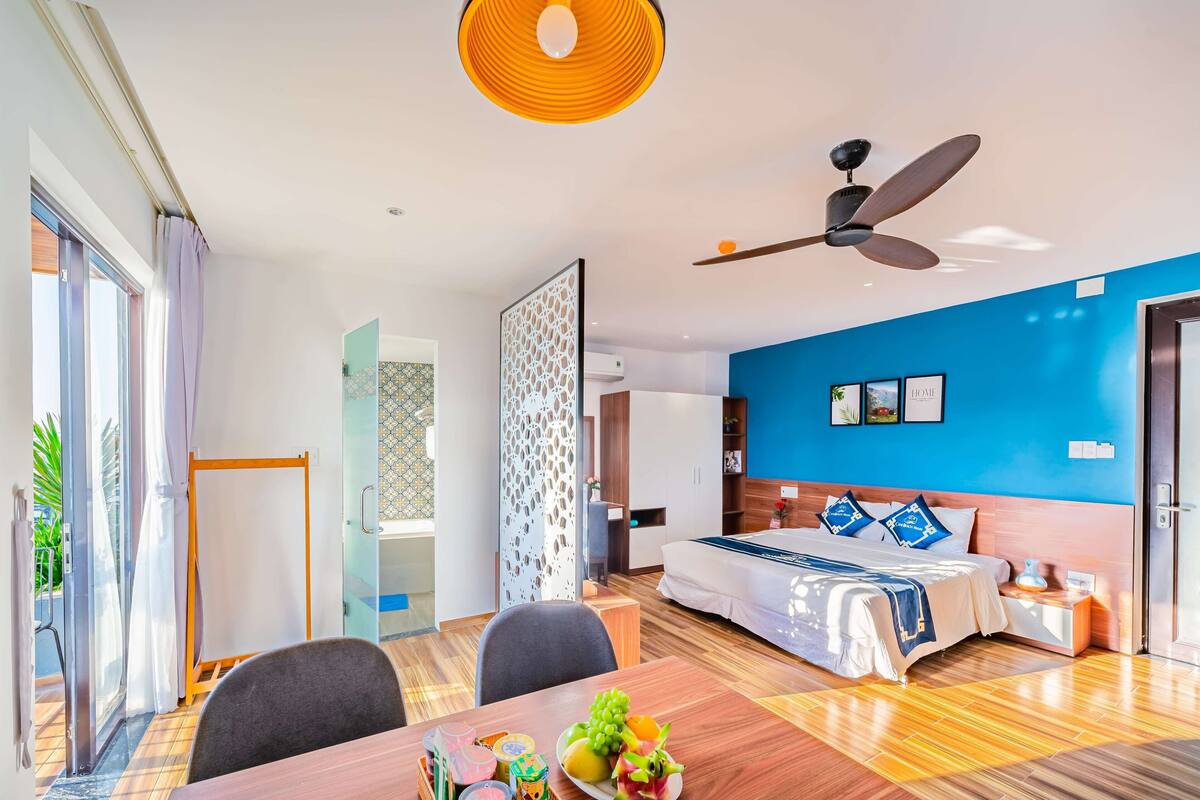 A modern hotel room with a large bed, a ceiling fan, a dining area with a table and chairs, and a view of the outside.