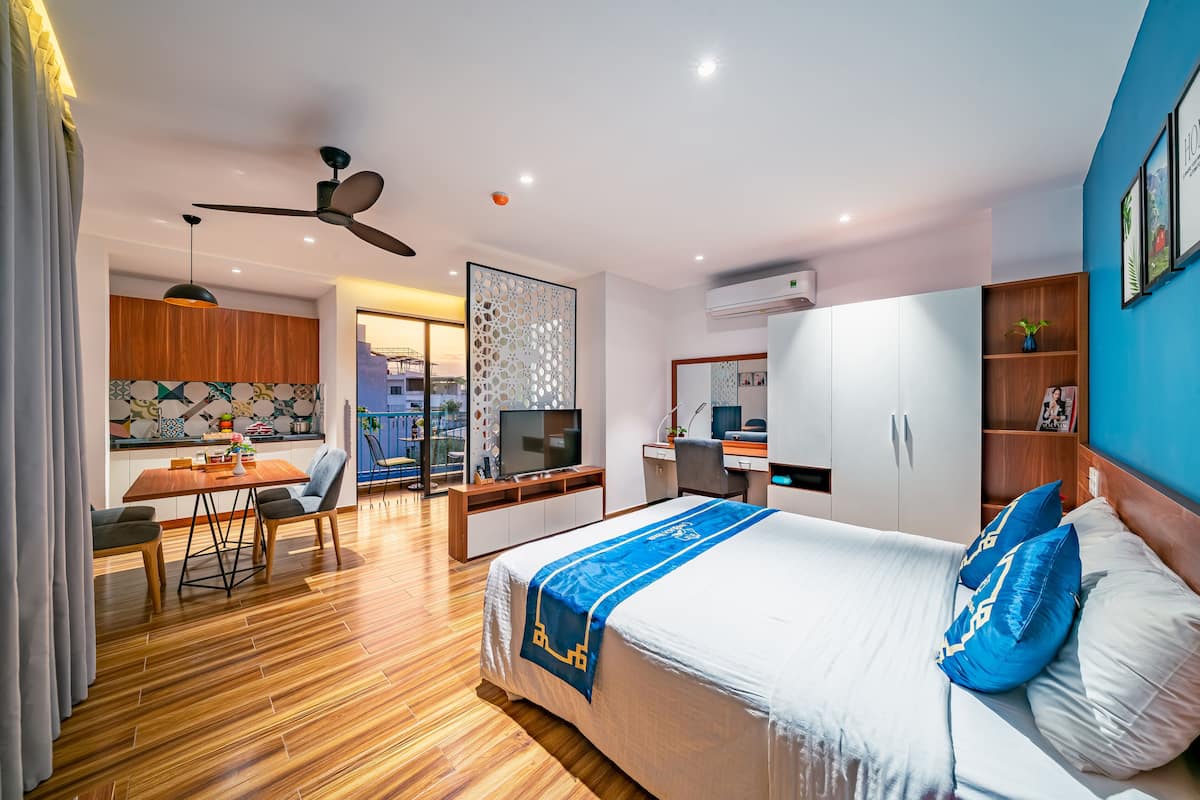 A modern hotel room with a bed, desk, and kitchenette.