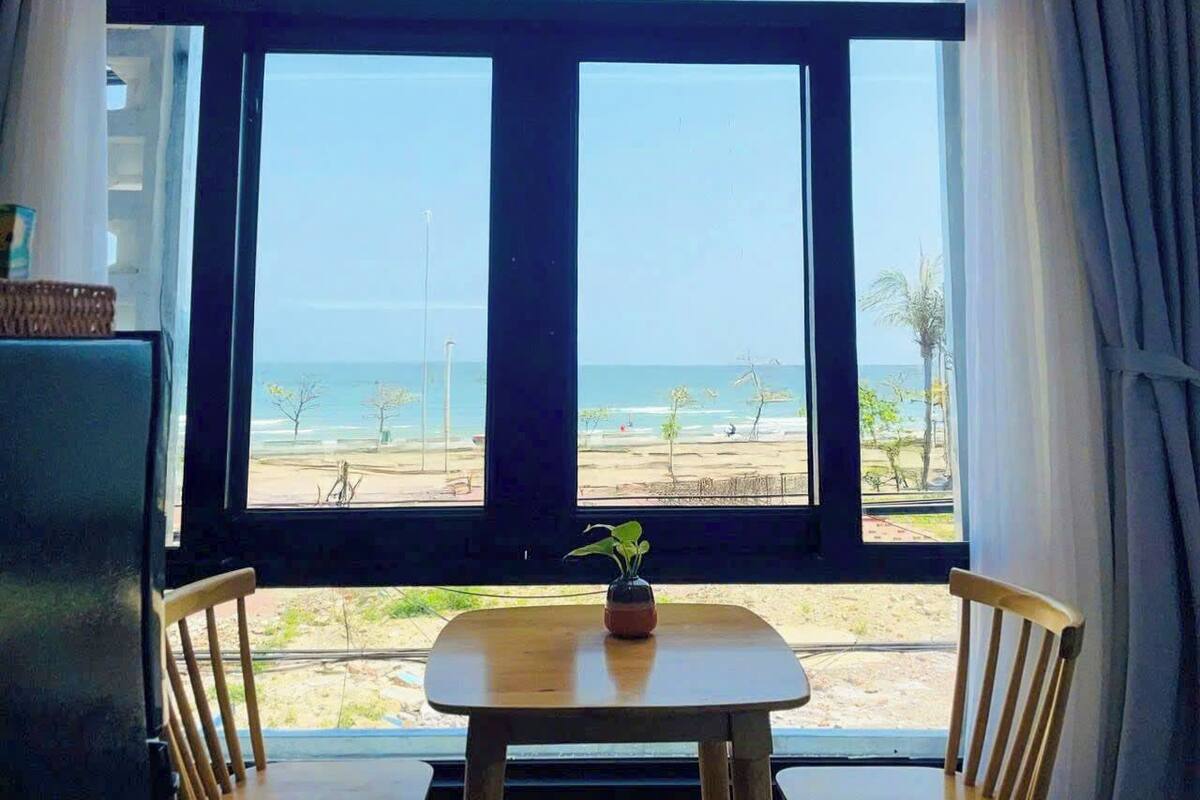 A room with a large window overlooking a beach, a small table with two chairs, and a potted plant on the table.