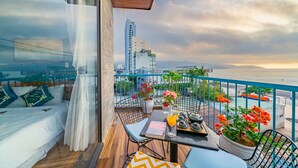 Un balcón con vistas al horizonte de la ciudad, una mesa con un libro y un vaso de jugo de naranja, y plantas en macetas con flores.