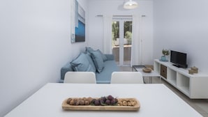 Un salon moderne avec un canapé blanc, une table à manger sur laquelle est disposé un plateau de fruits, un téléviseur posé sur un meuble blanc et une grande fenêtre agrémentée de rideaux.