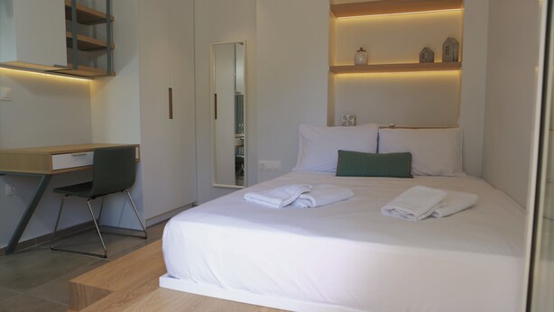 Une chambre d’hôtel avec un lit, un bureau et une chaise. Il y a un miroir au mur et des étagères au-dessus du lit.