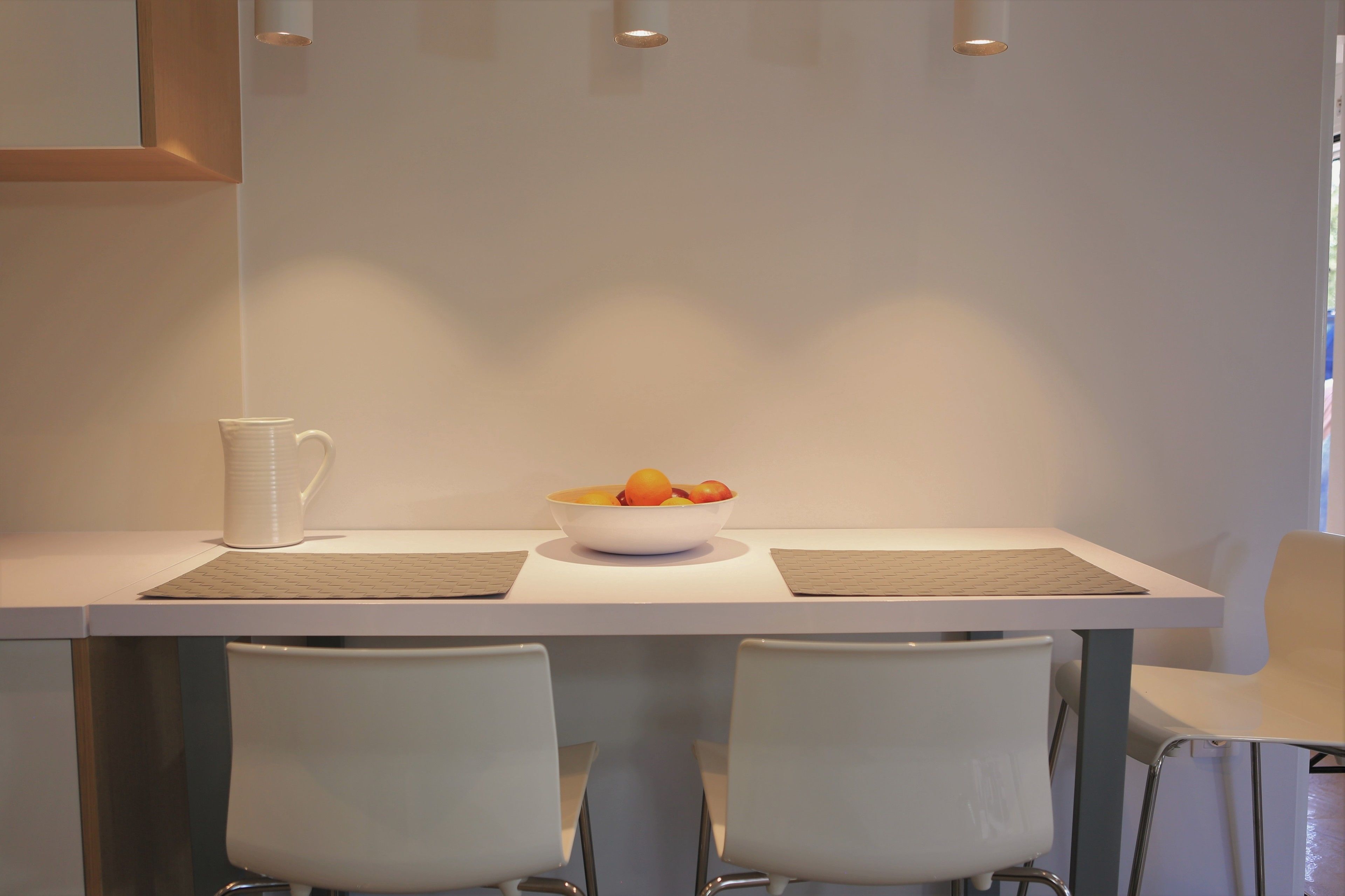 A modern dining area with a white table, two white chairs, a bowl of fruit, and a white pitcher.