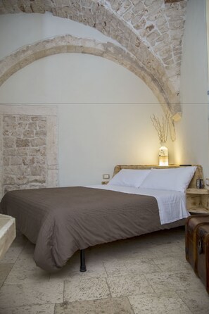 Un dormitorio con techo abovedado de piedra, una cama con colcha marrón, una mesita de noche con lámpara y una maleta en el suelo.