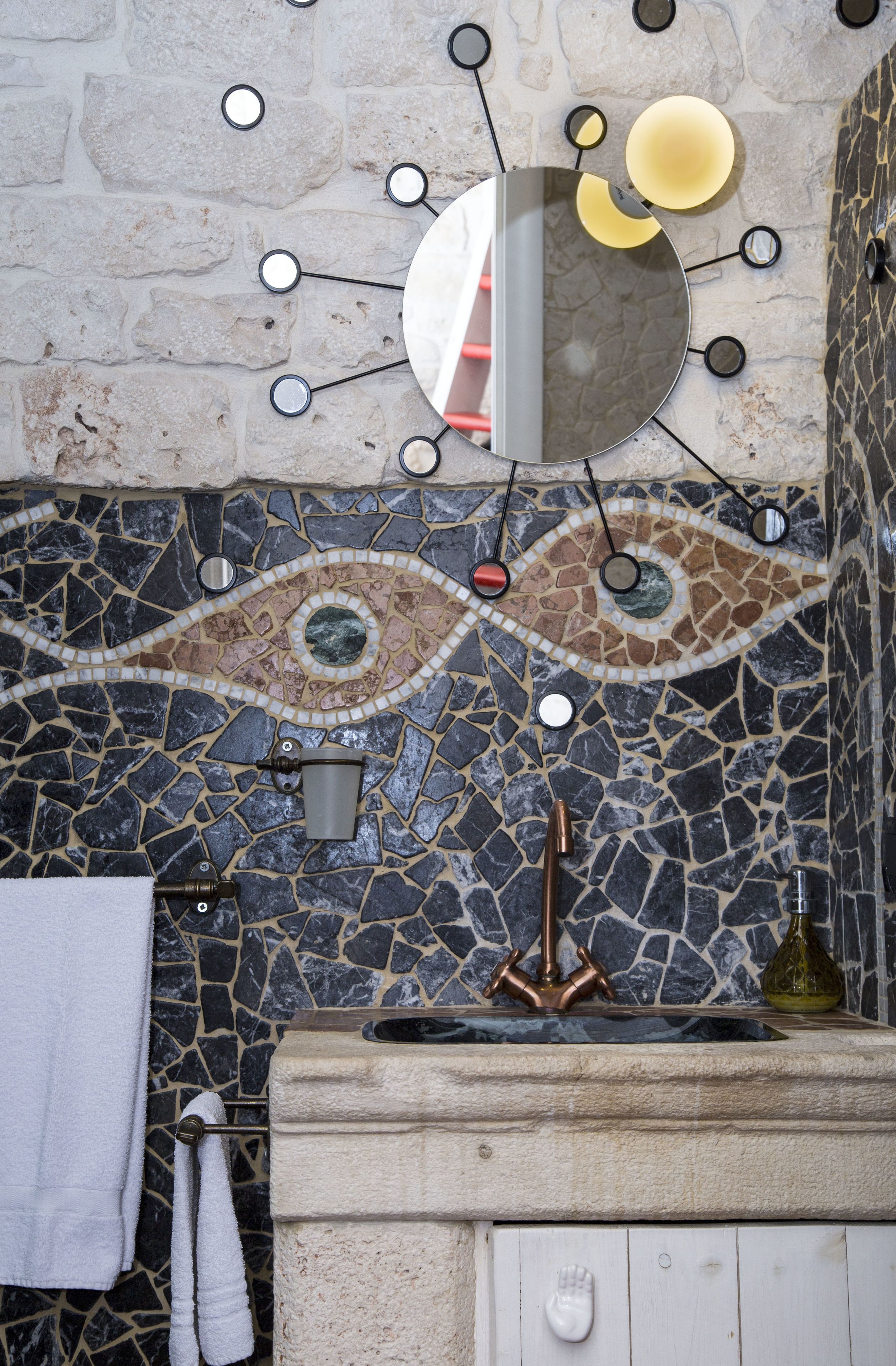 A bathroom with a mosaic tile backsplash, a mirror with circular lights, a towel rack, and a sink with a decorative bottle.