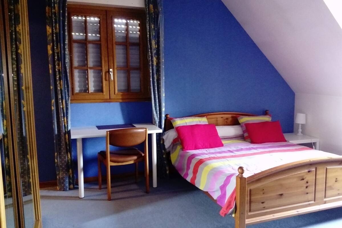 Un dormitorio con techo inclinado, una cama de madera con cabecera, un escritorio con silla y una ventana con cortinas.