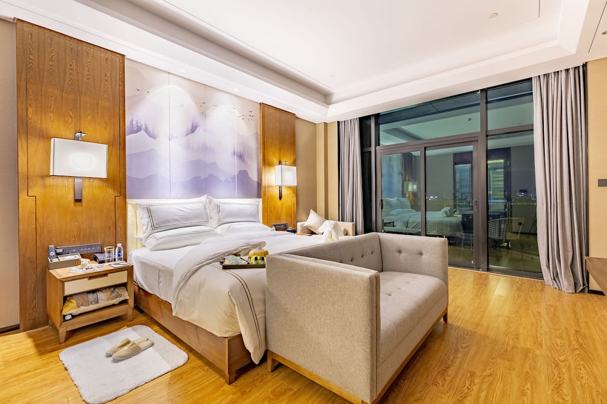 A hotel room with a bed, bedside table, lamp, and a view of the city through large windows.