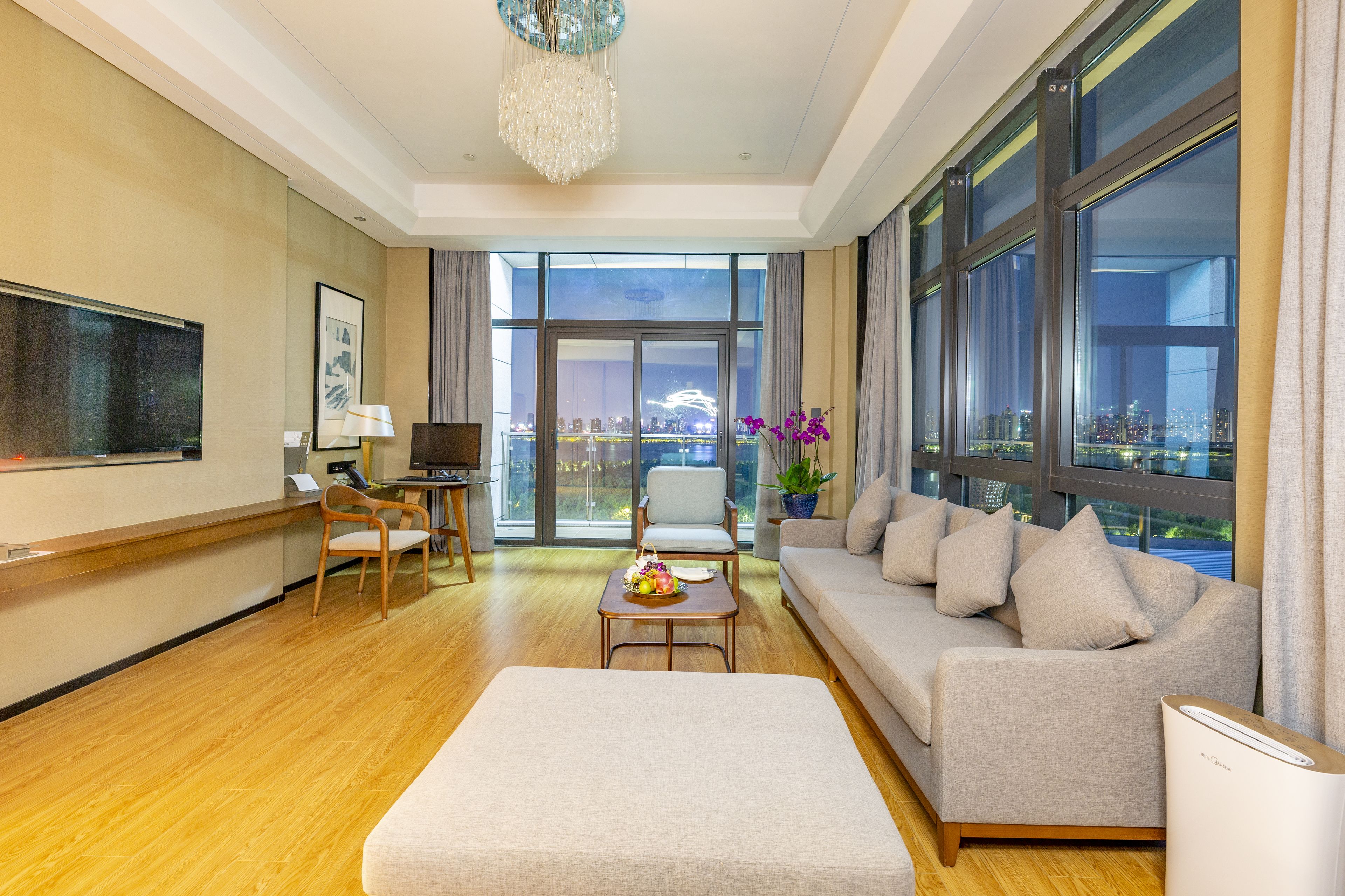 A modern hotel room with a large window, a sofa, a chair, a small table, and a TV.