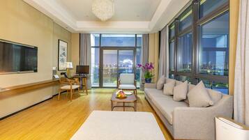 A modern hotel room with a large window, a sofa, a chair, a small table, and a TV.