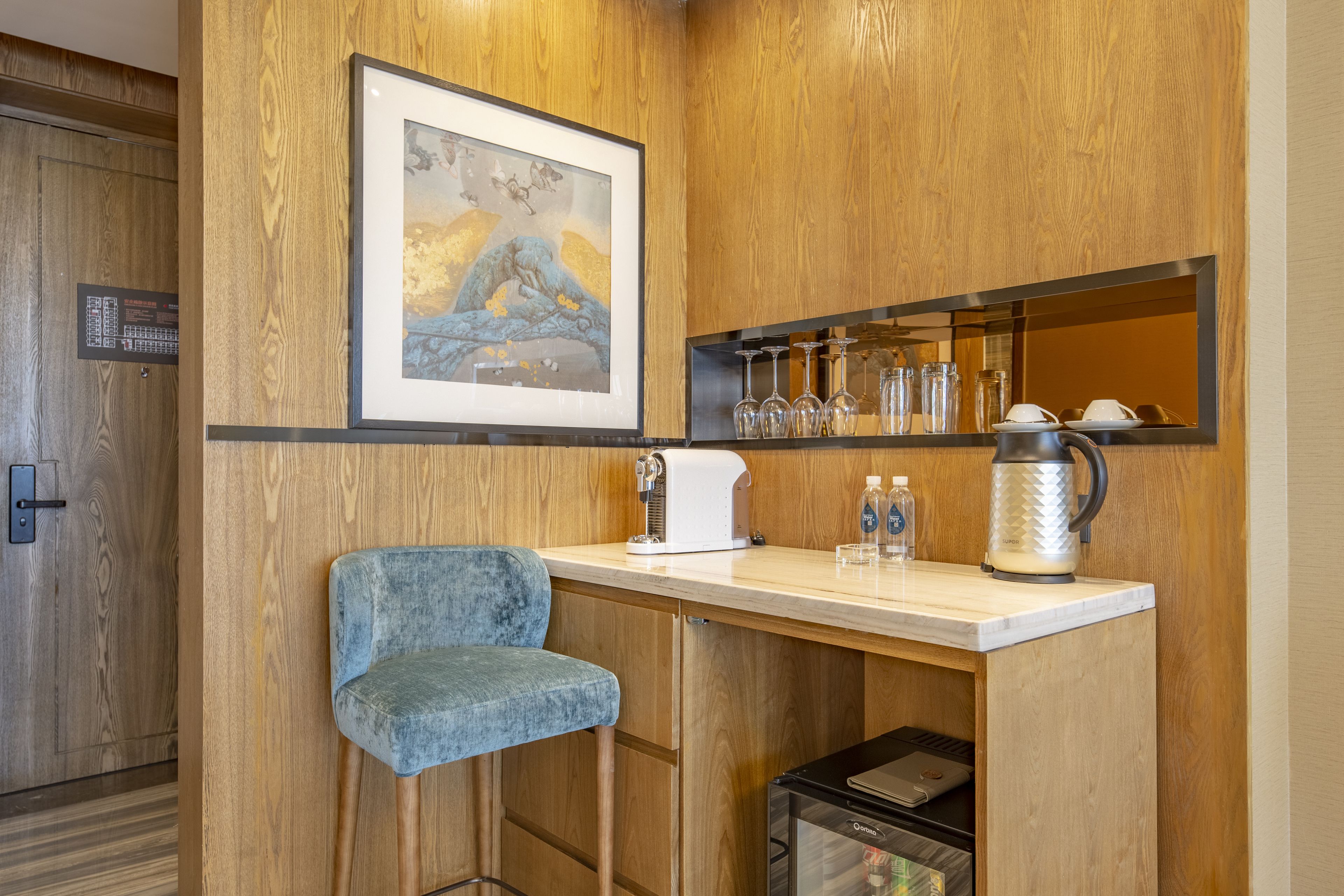 A bar area with a framed artwork, a bar stool, a coffee maker, and a shelf with glasses and bottles.