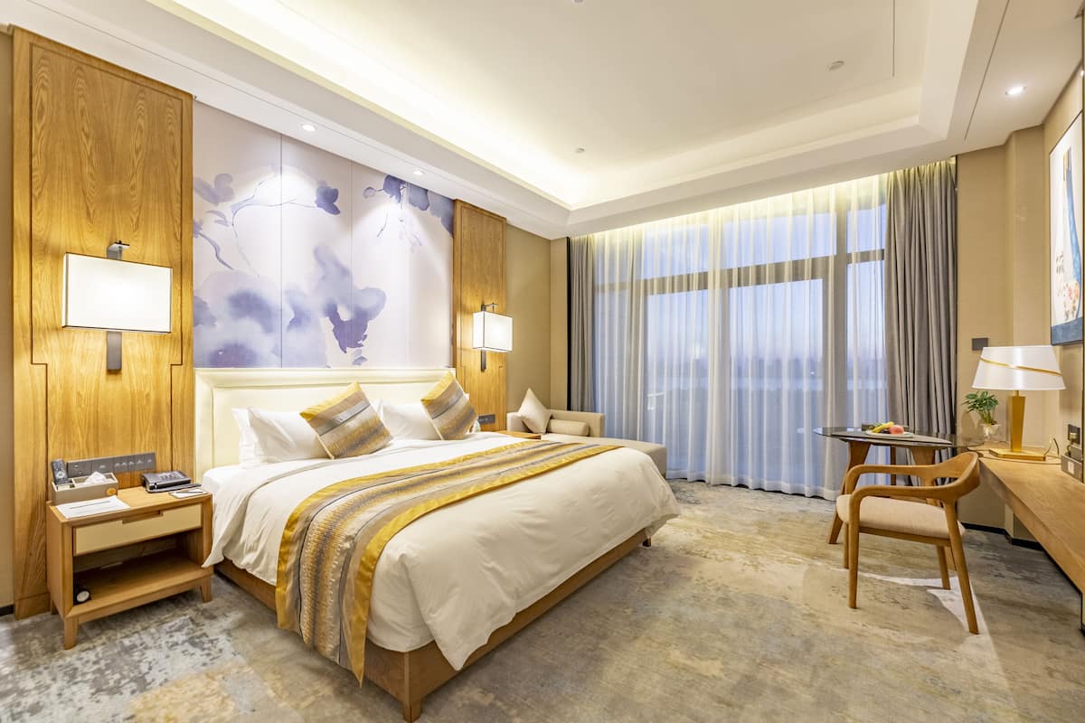 A hotel room with a large bed, wooden headboard, two bedside tables with lamps, a desk with a chair, and a large window with curtains.