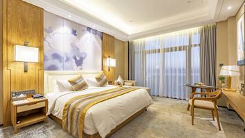 A hotel room with a large bed, wooden headboard, two bedside tables with lamps, a desk with a chair, and a large window with curtains.
