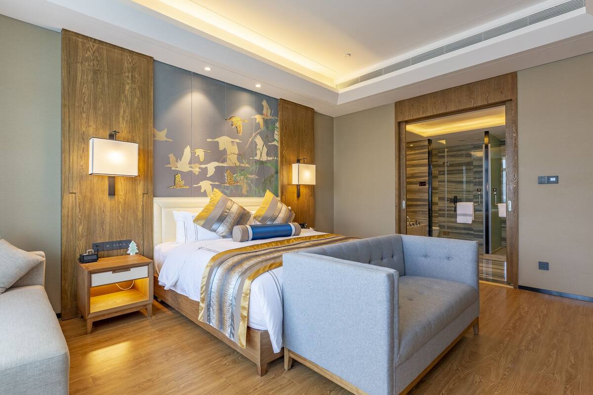 A modern hotel room with a large bed, a sofa, wooden paneling, and a mural on the wall.