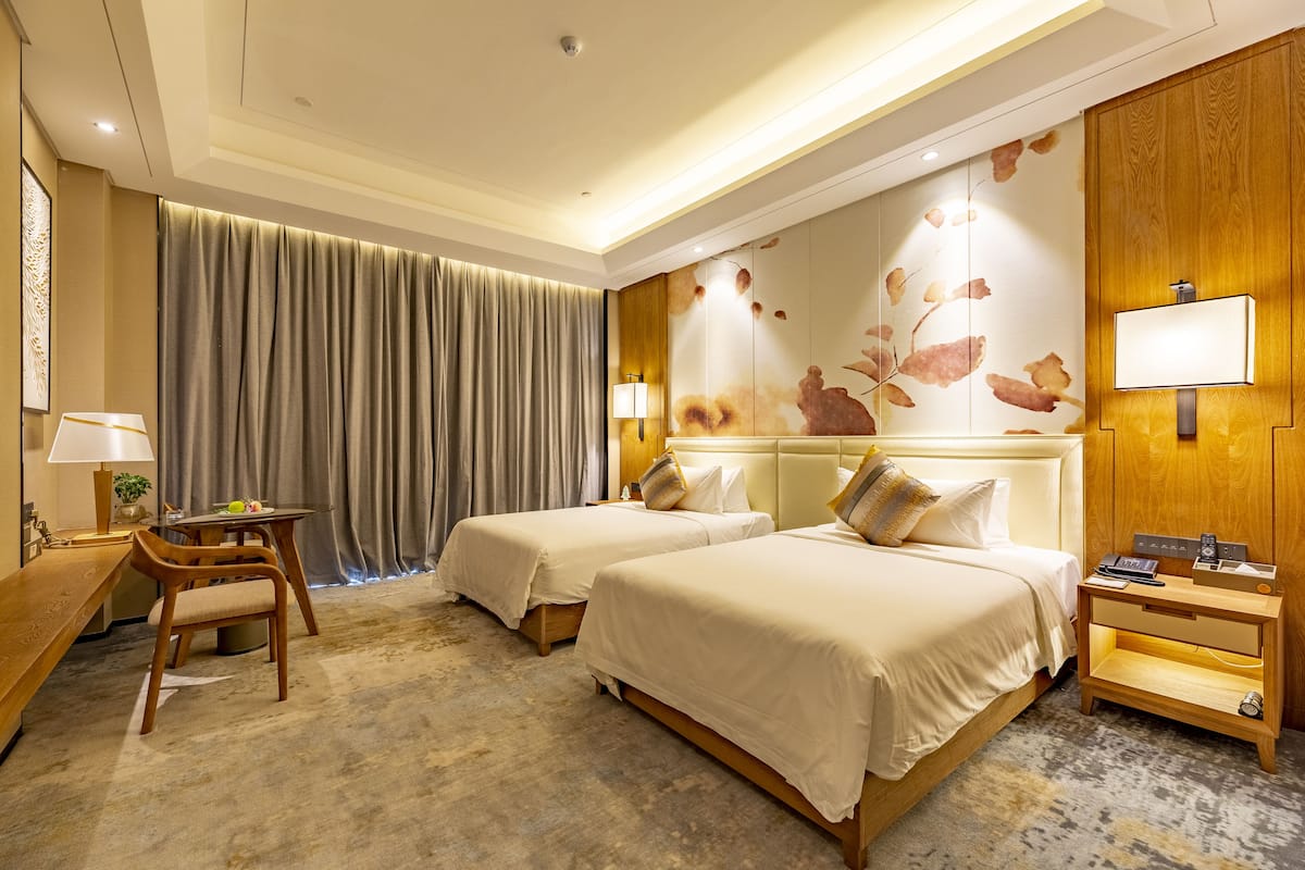A hotel room with two beds, a desk, a chair, and a nightstand with a lamp.