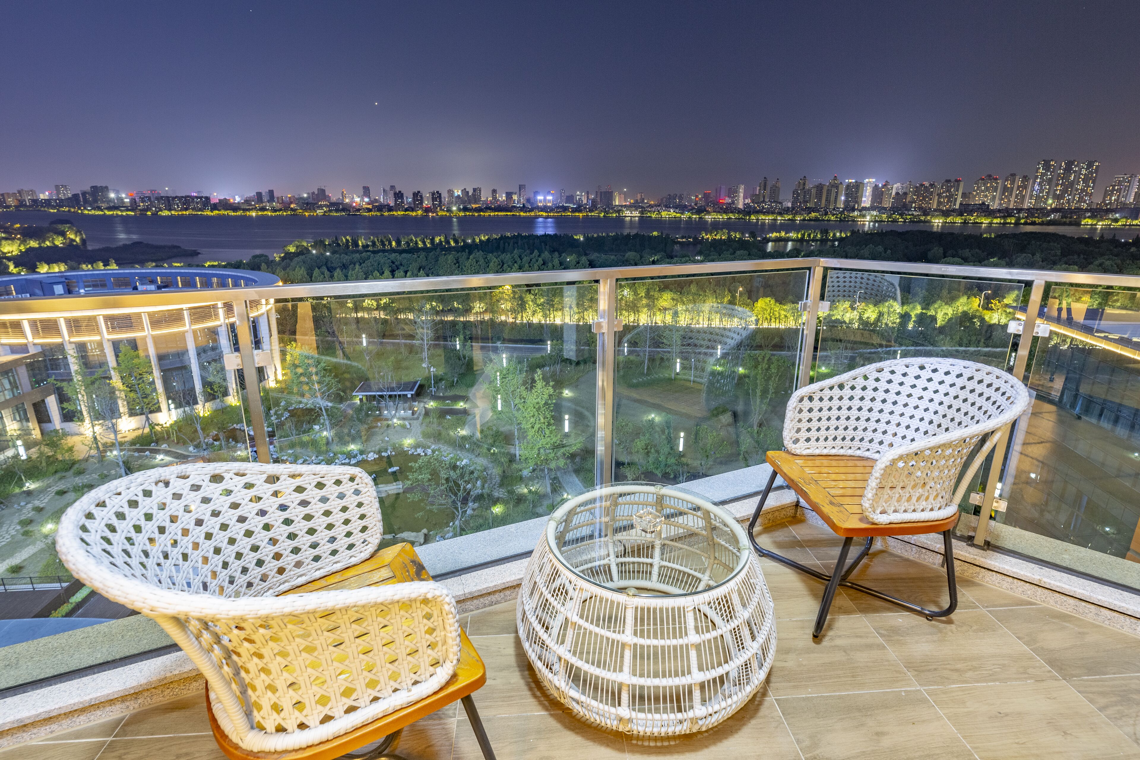 A balcony with two wicker chairs and a small round table, overlooking a cityscape with a river and illuminated buildings.