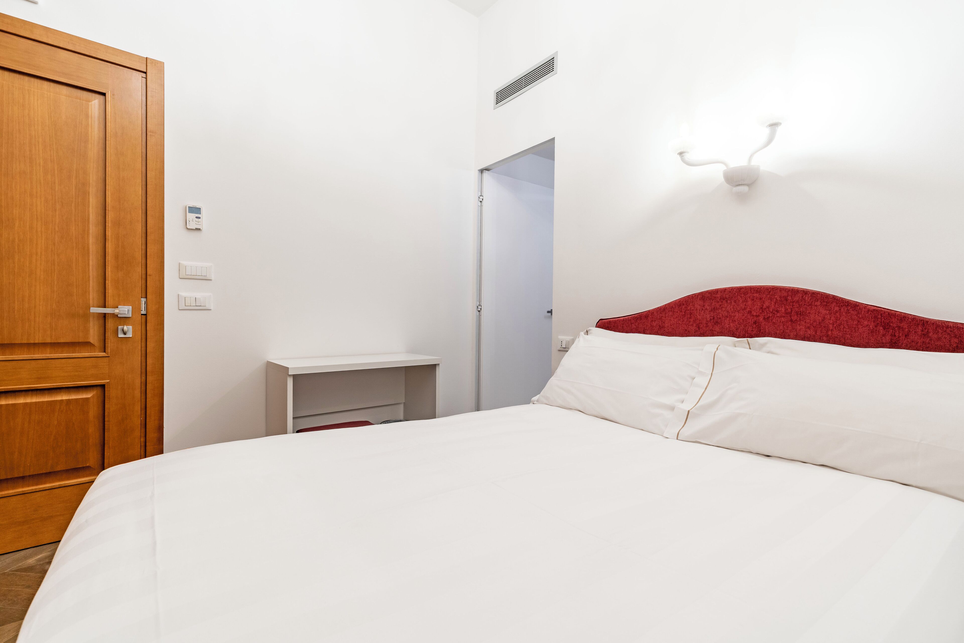 Habitación de hotel con puerta de madera, cama blanca con cabecera roja, mesita de noche y aplique de pared.