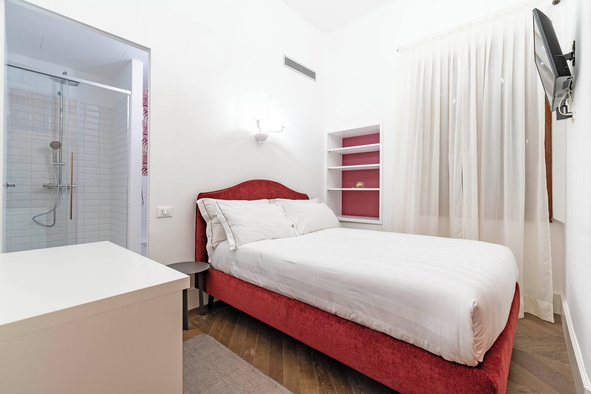 Une chambre avec un lit rouge, des murs blancs, une table de chevet blanche, une cabine de douche en verre et un rideau blanc.