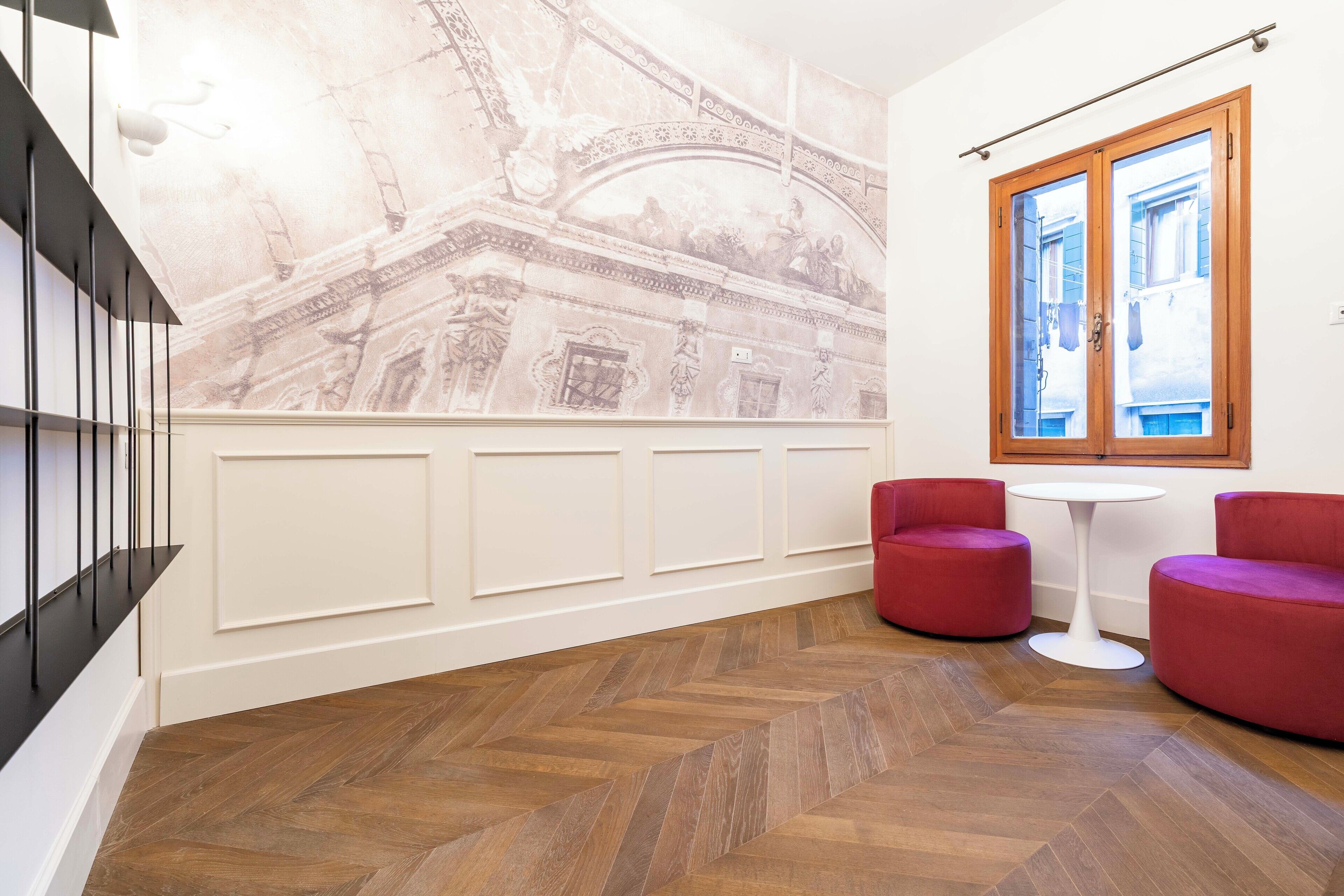 A hallway with a herringbone floor, a mural of a classical building, a wooden-framed window, and red upholstered chairs.