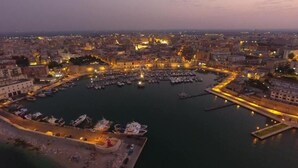 Vista aérea de uma paisagem urbana ao entardecer, com um porto repleto de barcos, cercado por edifícios e um calçadão à beira-mar.