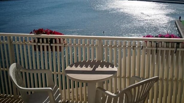 Uma varanda com uma mesa e duas cadeiras, dando vista para um mar calmo com um cais ao longe.