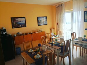 A dining area with wooden chairs and a table set for four, a small kitchen area with a refrigerator and cabinets, and two framed pictures on the wall.