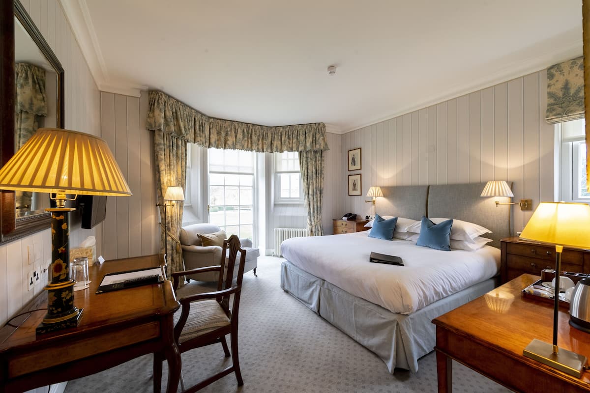 A hotel room with a large bed, two bedside tables, a desk with a lamp, a chair, and a window with curtains.