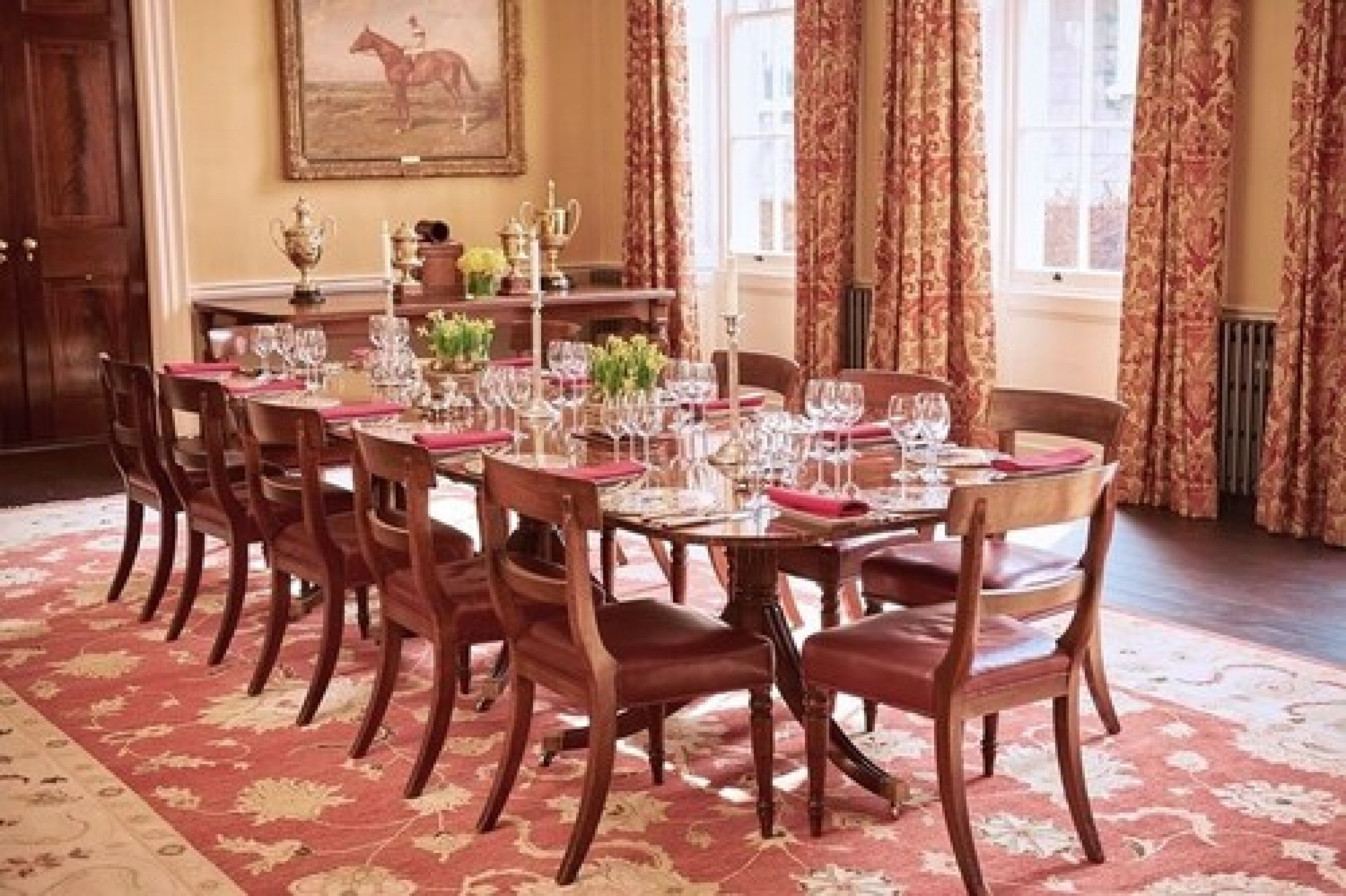 A formal dining room with a long table set for multiple guests, featuring wooden chairs and a patterned carpet.