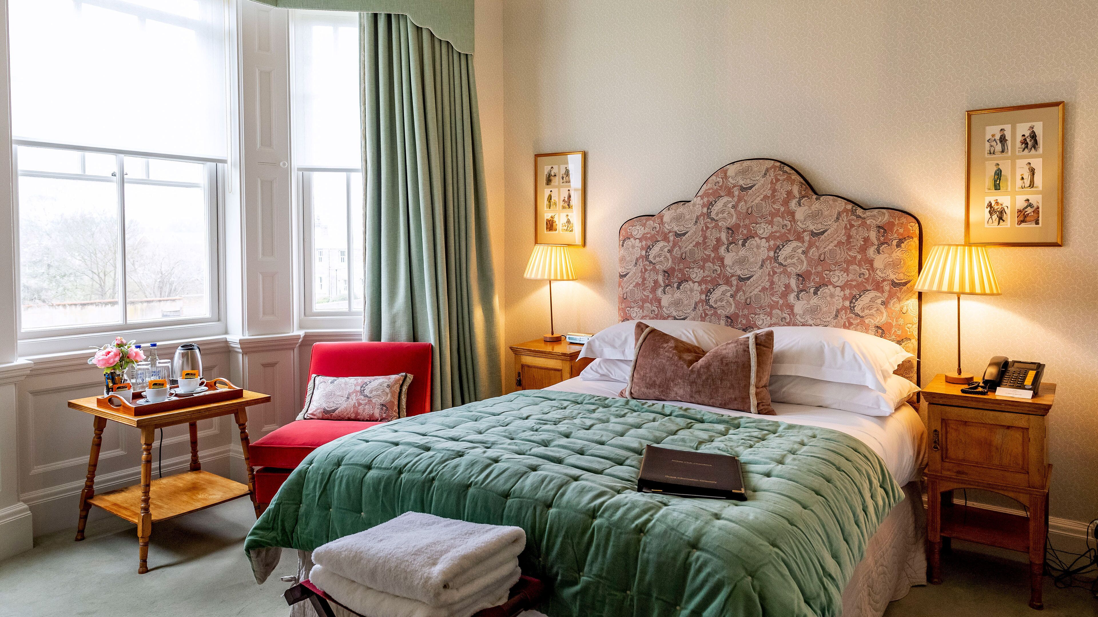 A hotel room with a large bed, a chair, a bedside table with a lamp, a bedside phone, and a window with curtains.