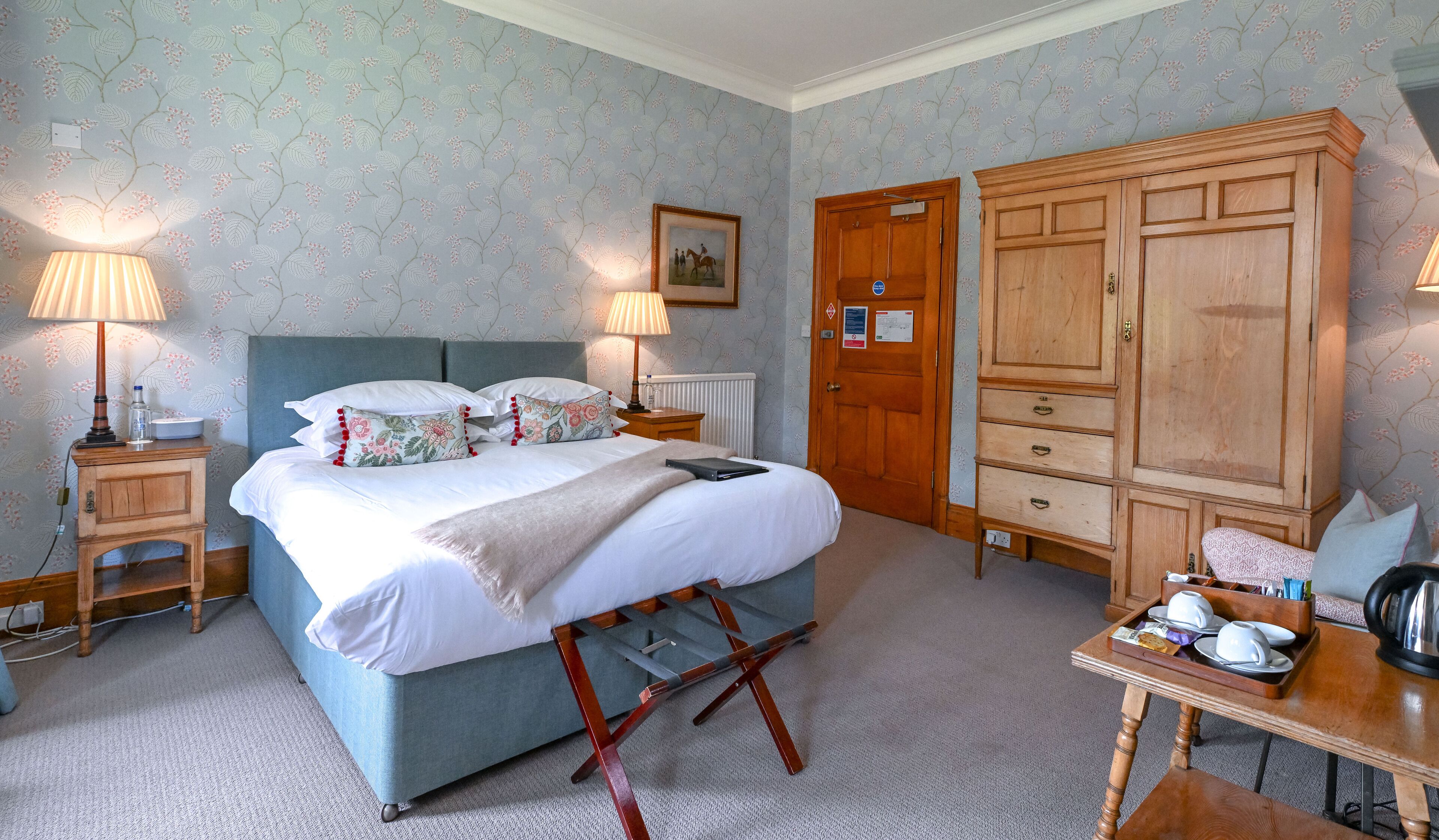 A bedroom with a bed, bedside lamps, a wooden wardrobe, a small table with cups and a kettle, and a radiator.