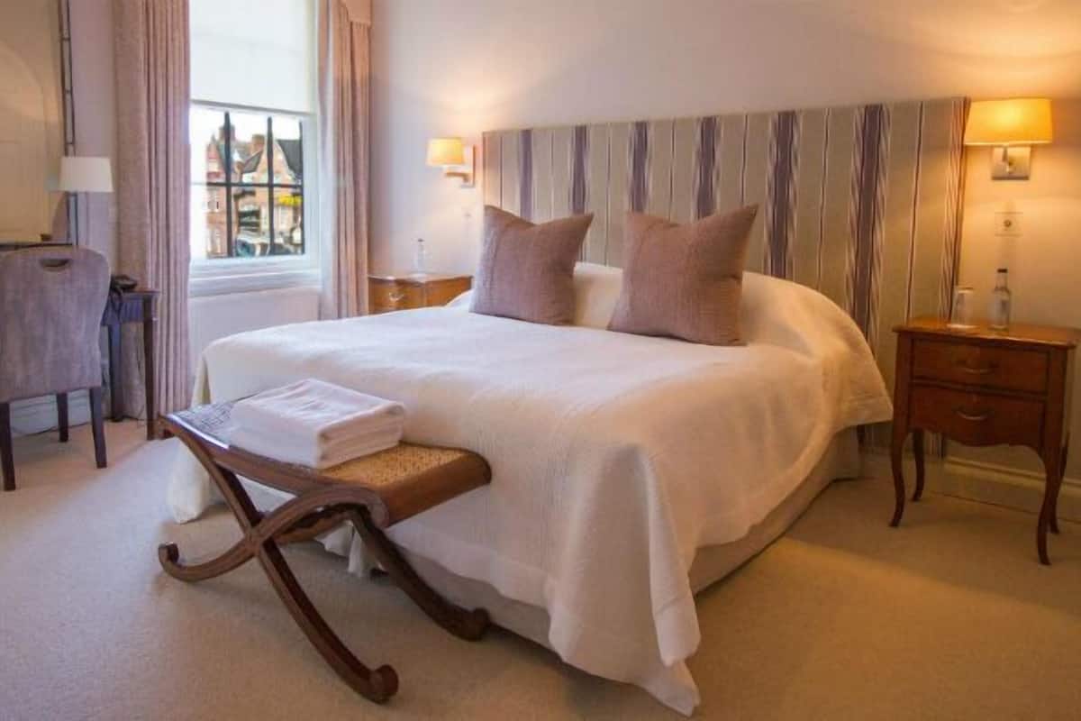 A hotel room with a large bed, a desk with a chair, a small table with a lamp, and a window with curtains.