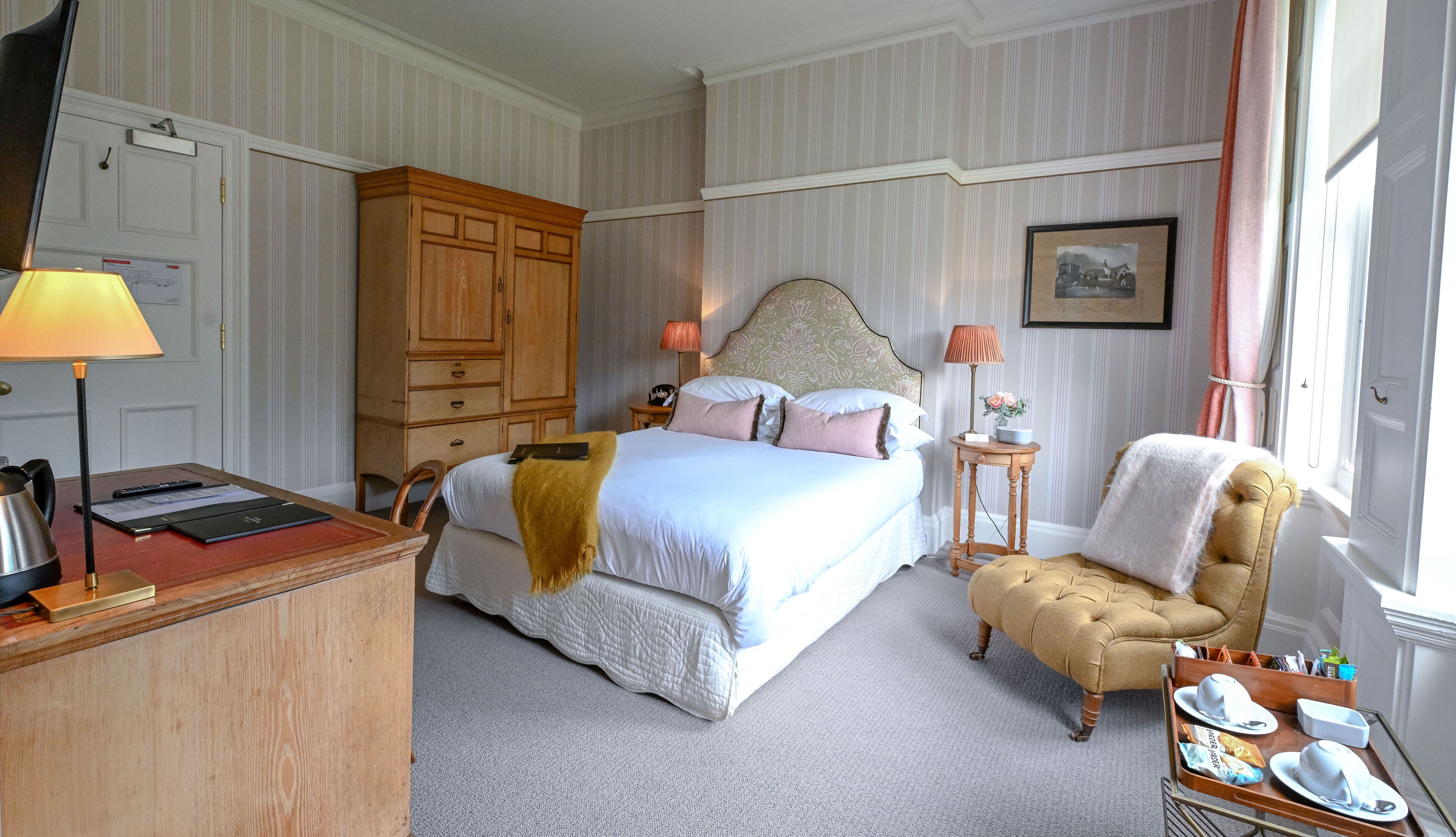 Habitación de hotel con cama, escritorio con lámpara, silla, mesita de noche con lámpara y ventana con cortinas.