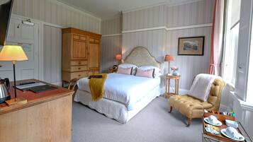 Quarto de hotel com cama, escrivaninha com luminária, cadeira, mesa de apoio com luminária e janela com cortinas.