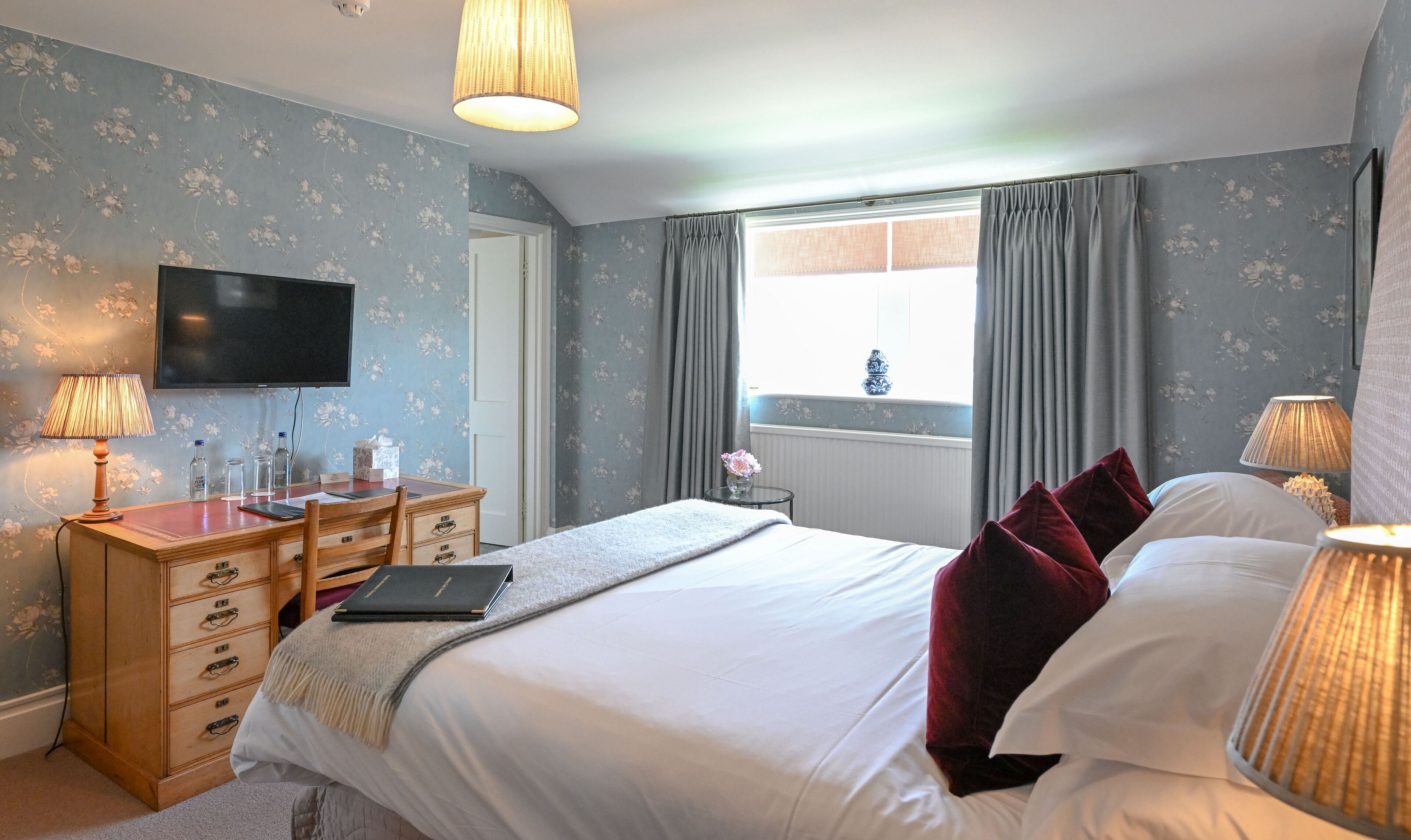 A hotel bedroom with a bed, bedside lamps, a desk with a laptop, a dresser, and a TV.