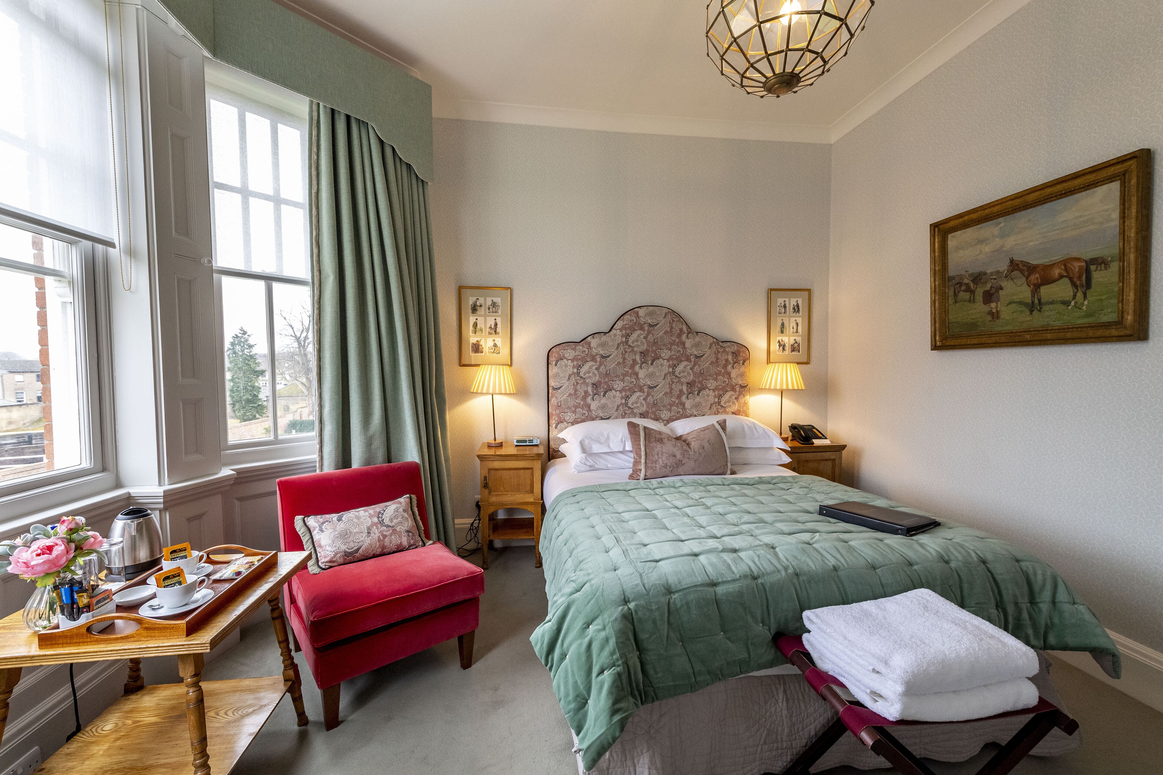 A hotel room with a large bed, a red armchair, a wooden side table with a tray of breakfast items, and a painting on the wall.