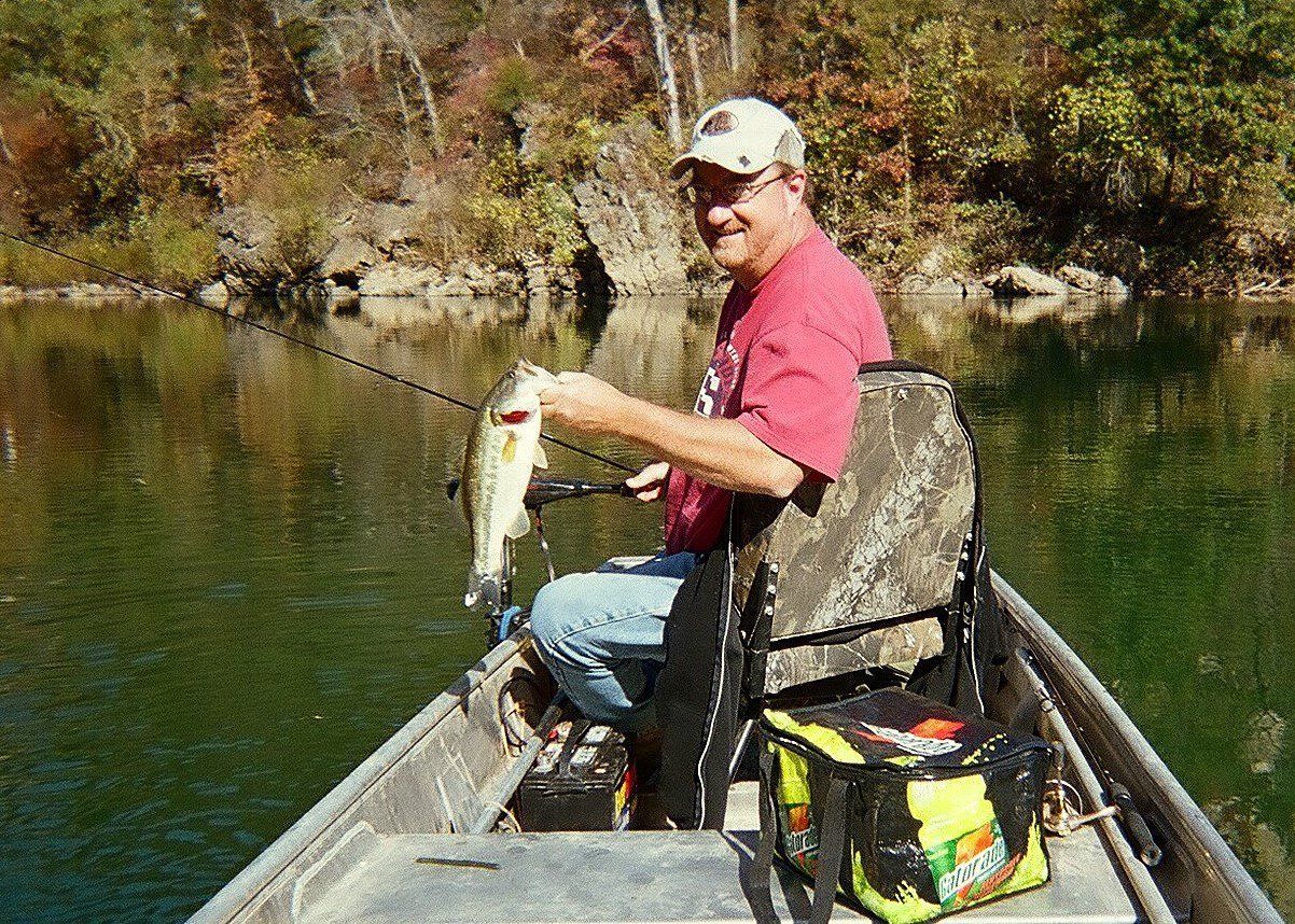 A person in a boat holding a fish, with a bag of bait and a tackle box visible.