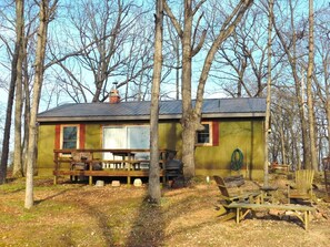 A rustic cabin with a wooden deck, a barbecue grill, and a large window.