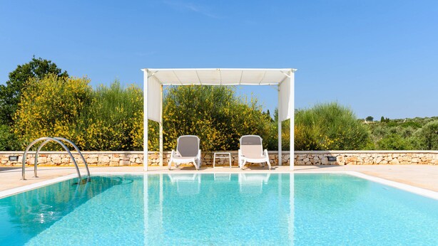 Ein klares Schwimmbecken mit zwei Liegestühlen unter einer weißen Pergola, umgeben von viel Grün.