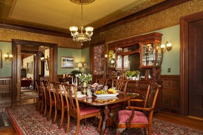 Formal dining room with ornate table, decorative ceiling, and wall paneling, featuring a chandelier and sconces.