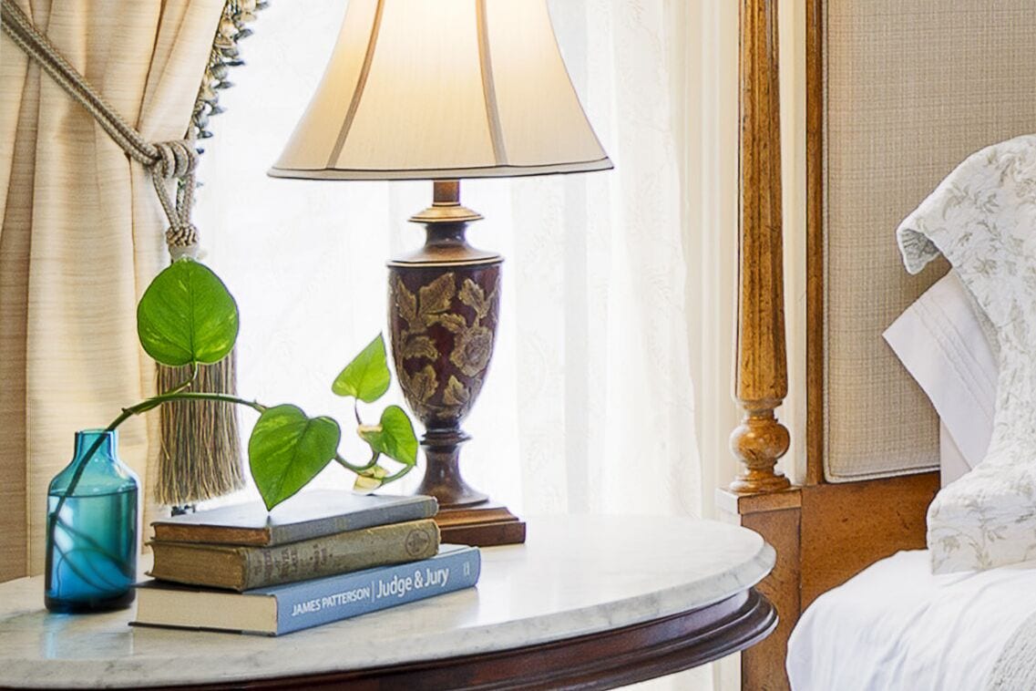 A neatly arranged bedside table with a lamp, a book titled 'Judge & Jury' by James Patterson, a blue vase with a plant, and a bed with a white floral bedspread.