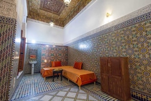 Ein Zimmer mit Schachbrettmuster auf dem Boden, einem Bett mit orangefarbener Bettwäsche, einem Holzschrank und einer Wand mit aufwendigen Fliesenmustern.