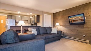 A modern living room with a grey sofa, a TV mounted on a wooden wall, and a kitchen in the background. - Storm Meadows 300-400 at Christie Base (Steamboat Springs)