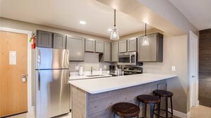 A modern kitchen with a stainless steel refrigerator, a microwave, and a built-in oven. There are three bar stools and pendant lights above the island. - Storm Meadows 300-400 at Christie Base (Steamboat Springs)