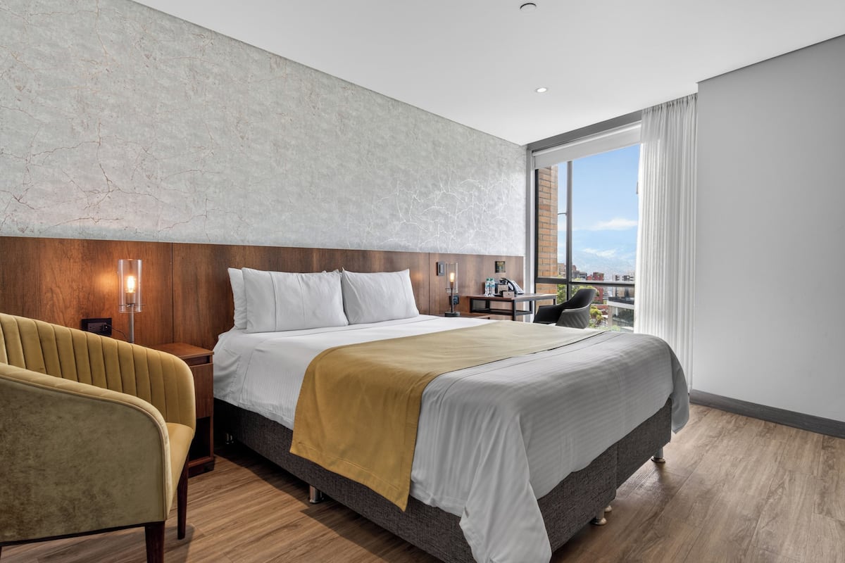 A modern hotel room with a large bed, a wooden headboard, a bedside table with a lamp, a chair, and a view of the city through a large window.