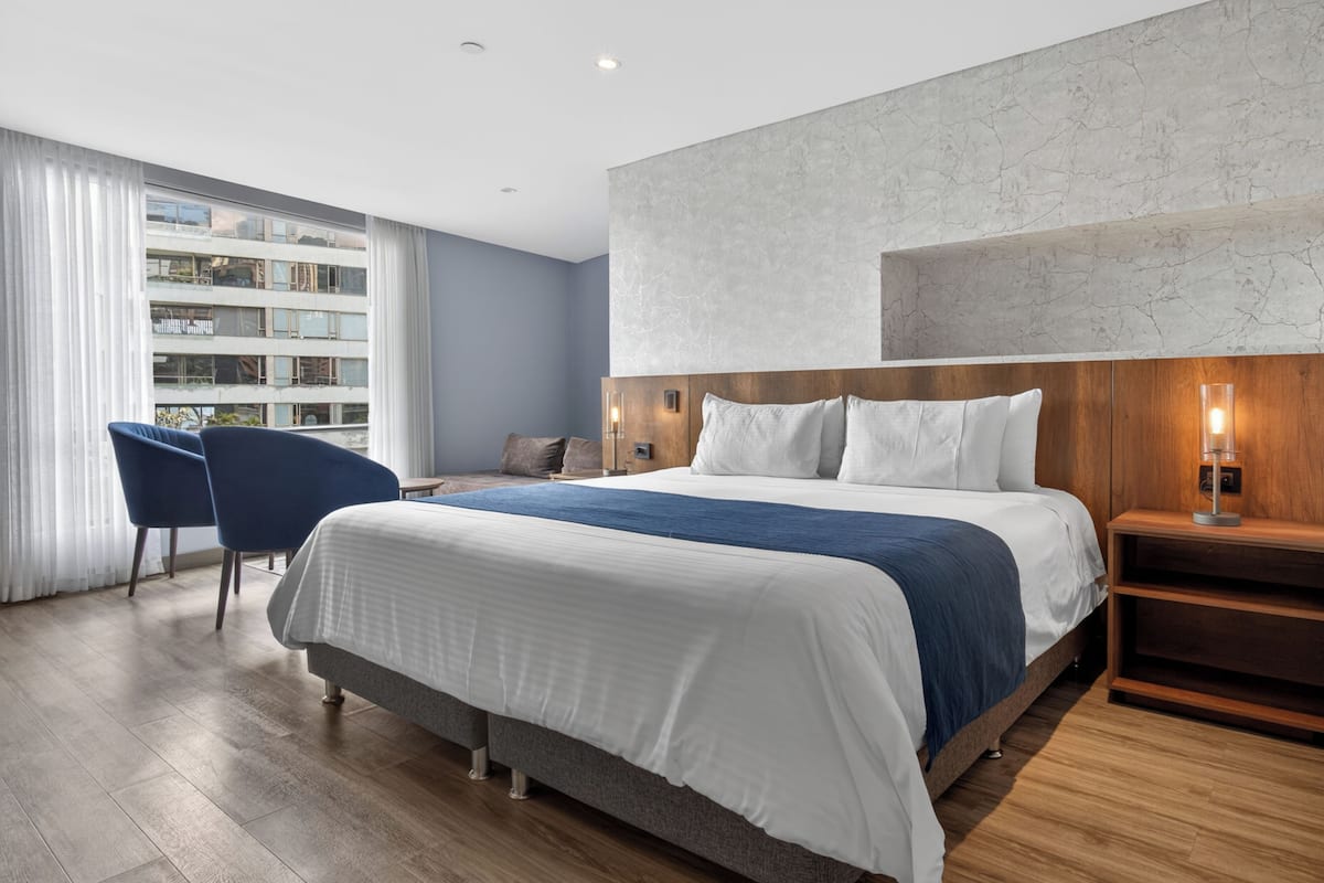 A modern hotel room with a large bed, a blue chair, a wooden nightstand, and a view of a building through the window.