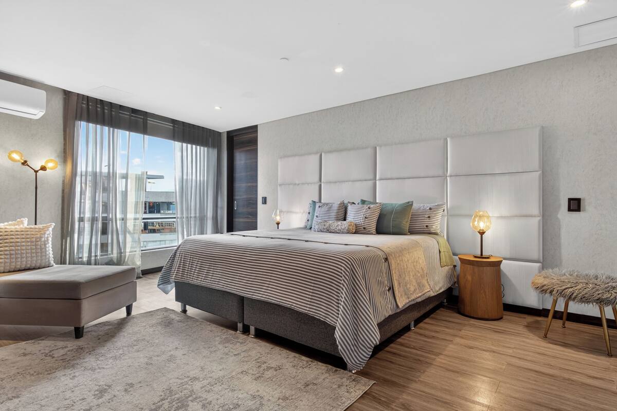 A modern bedroom with a large bed, a side table, a chair, and a view of the city.